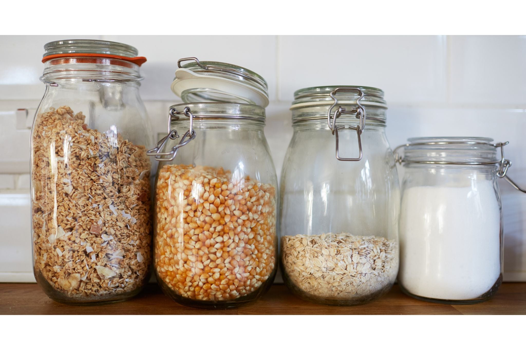 Vier Glasbehälter mit verschiedenen Lebensmitteln: Müsli, Popcornmais, Haferflocken und Zucker, auf einer Küchenarbeitsplatte